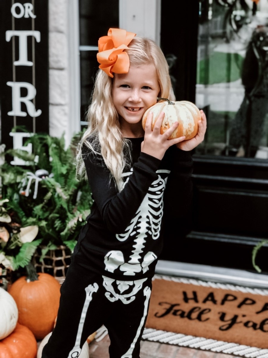 Halloween Treats | Easy Halloween Activities for Families by popular Houston lifestyle blog, Haute and Humid: image of a little girl holding pumpkins on a front porch with a Target Project 62 Chevron Woven Area Rug, Etsy Happy Fall Yall Welcome Doormat, and wearing Amazon Little Pajamas skeleton glow-in-the-dark pajama sets.