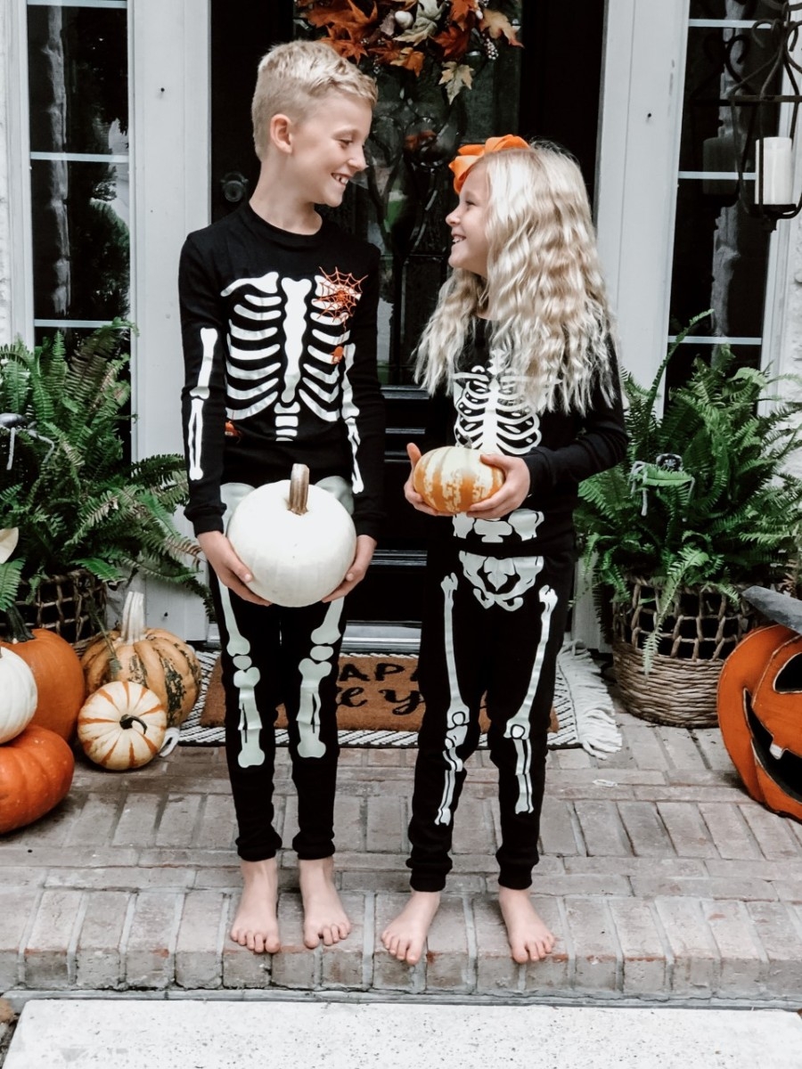 kids Halloween pajamas | Easy Halloween Activities for Families by popular Houston lifestyle blog, Haute and Humid: image of two kids holding pumpkins on a front porch with a Target Project 62 Chevron Woven Area Rug, Etsy Happy Fall Yall Welcome Doormat, and wearing Amazon Little Pajamas skeleton glow-in-the-dark pajama sets. 