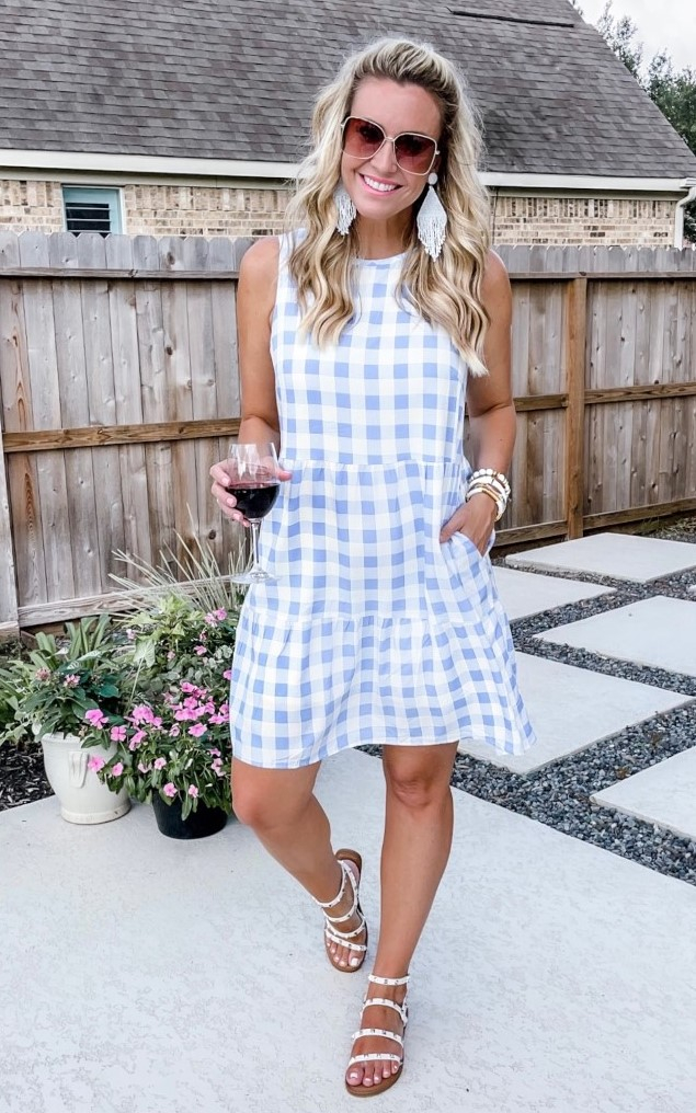 amazon prime dress | Amazon Prime Day by popular Houston fashion blog, Haute and Humid: image of a woman standing outside and holding a glass of red wine and wearing a blue and white buffalo check tiered sleeveless mini dress with white strap sandals, white bead statement earrings and sunglasses. 