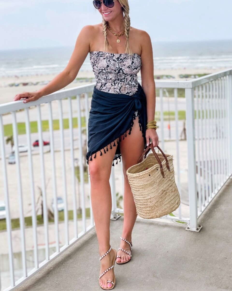 amazon one piece swimsuit | Amazon Prime Day by popular Houston fashion blog, Haute and Humid: image of a woman standing outside on a balcony and wearing a strapless one piece snake print swimsuit and black tassel Sarang with sunglasses, pearl studded sandals, and holding a straw tote bag.