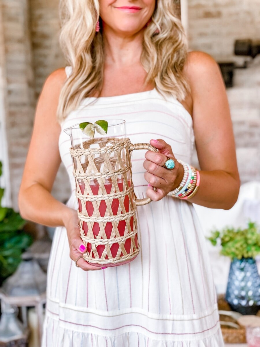 summer cocktails | Pitcher Cocktails by popular Houston lifestyle blog, Haute and Humid: image of a woman wearing a white stripe maxi dress, pink statement earrings, sunglasses, tan gold studded cross strap slide sandals and holding a glass pitcher covered in raffia that's filled with a pink cocktail drink. 