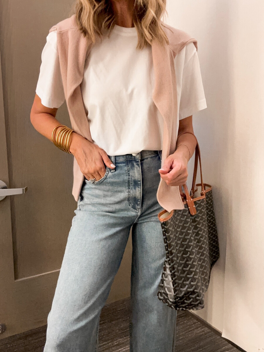 Woman styling a blush pink sweater draped over a white T shirt with light wash jeans and a designer tote bag.
